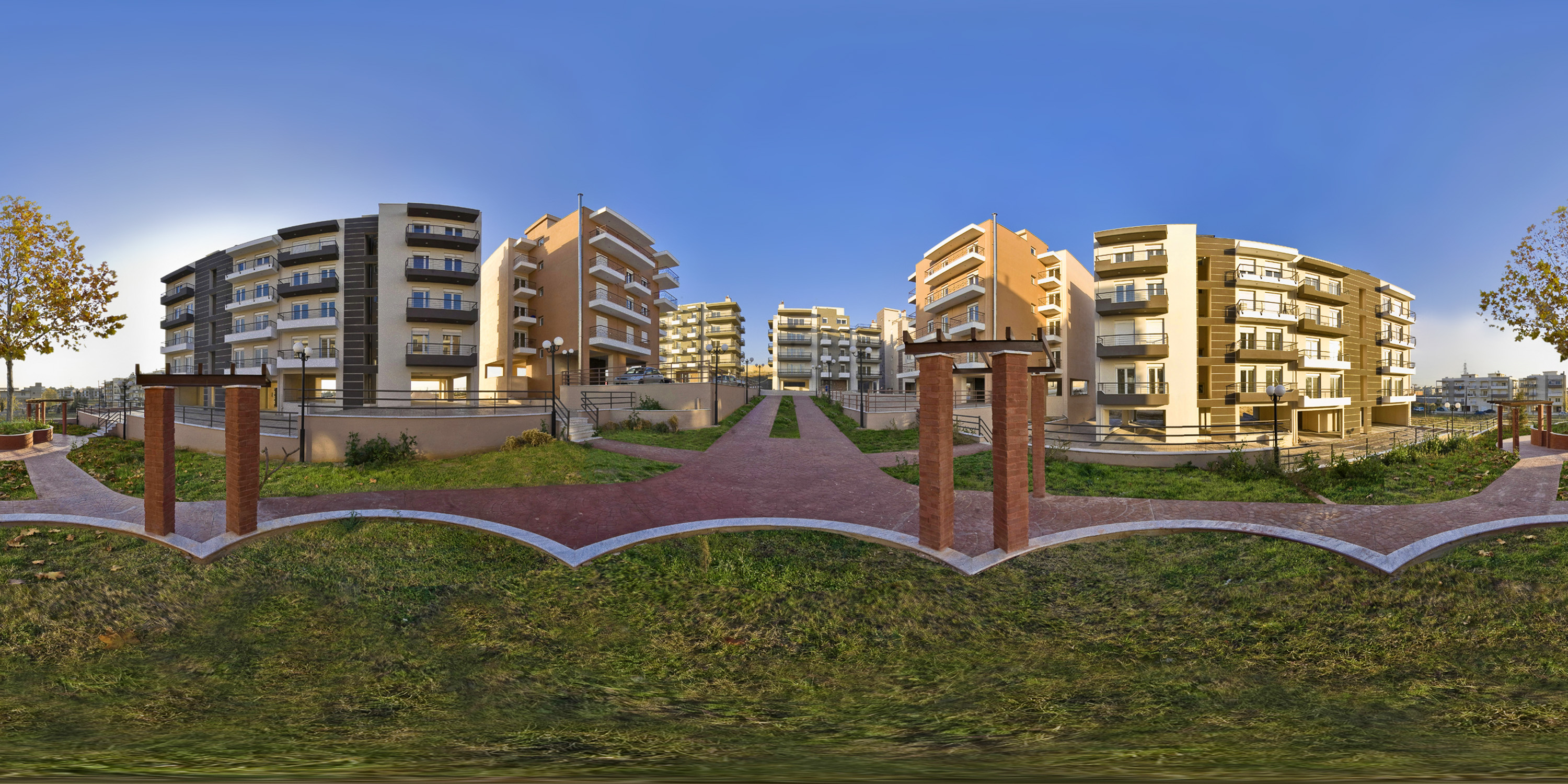 An equirectangular 360-degree photosphere of a modern residential apartment complex with multi-story buildings, manicured lawns, paved walkways, and brick pergolas under a clear blue sky.